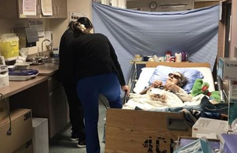 An elderly woman, lying in a hospital bed, wearing sunglasses, in a cluttered room with two health-care workers at her bedside.