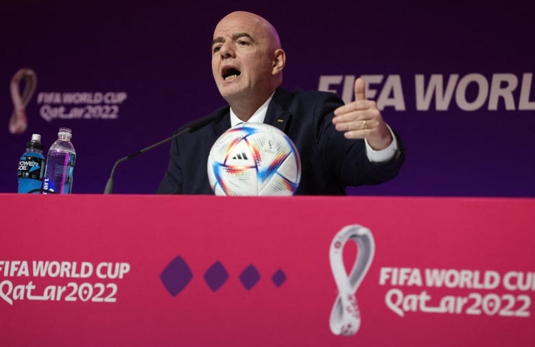 A bald man wearing a suit gestures while seated at a table with a soccer ball on it.