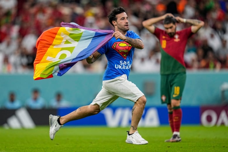 A tattooed man with brown hair wearing a blue T-shirt that has a Superman logo and text on it, and holding a rainbow flag, runs on the grass of a soccer pitch while a soccer player in a red and green uniform looks on.