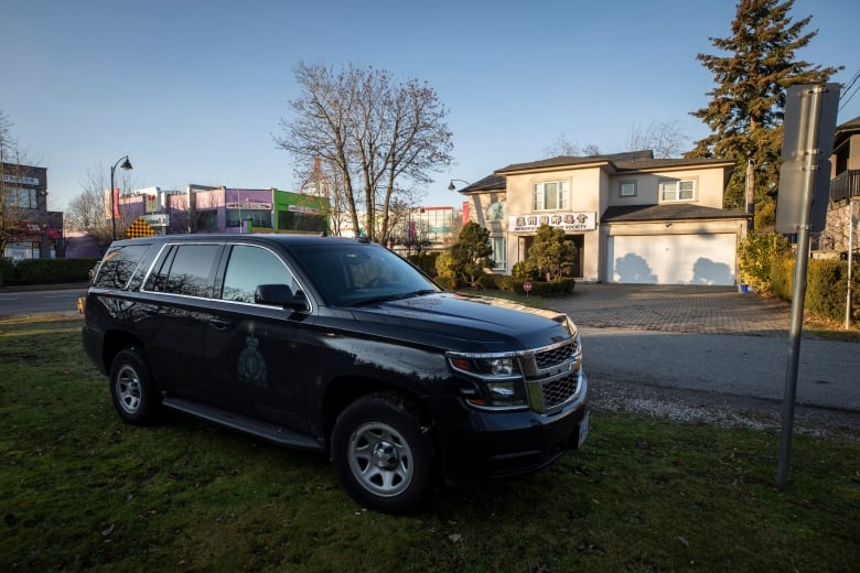 A black SUV with the RCMP logo on its side is shown in the foreground at an angle to the camera. In the background is a beige, house-like building with a sign in English and Chinese characters.