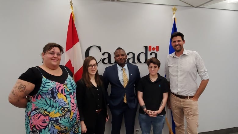 Five people stand in a line in front of a Canadian flag and a "Canada" sign on a white wall.