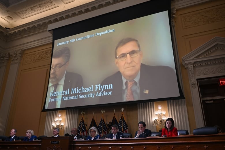 Two men in suit jackets and ties are shown on a video screen above and behind a panel of legislators that includes men and women.