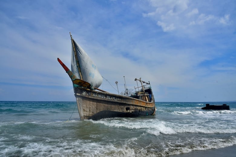 A wooden sail boat idles in the water a few feet from shore.