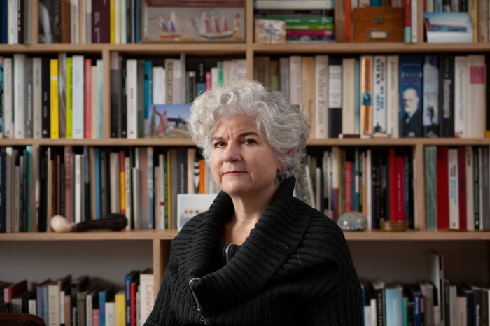 An older woman with short wavy grey hair wearing a big black jumper looks serious in front of bookshelves