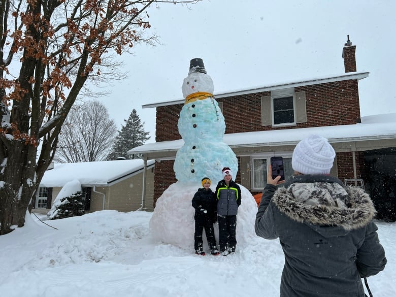 A woman takes a photo of two young boys standing in front of a snowman.