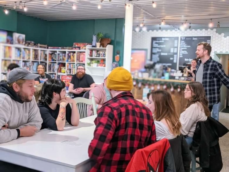 A group of people sit at tables at a board game café, listening to the store owner. 