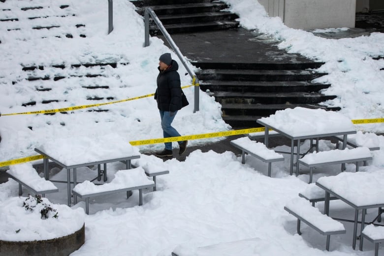 A person in a coat and jeans walks along a cleared path through piles of snow. There are picnic tables covered in snow, with yellow caution tape separating them from the person.