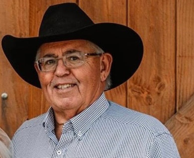 A man wearing a black cowboy hat and a blue-and-white-checkered shirt smiles at the camera.