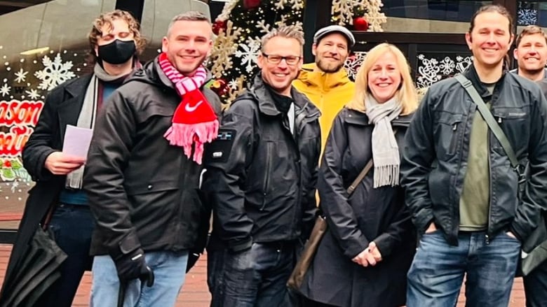 A group of people framed by Christmas decorations and dressed in coats and festive scarves smile for the camera, including the person in the centre, who is wearing a thin blue line patch on his right shoulder.