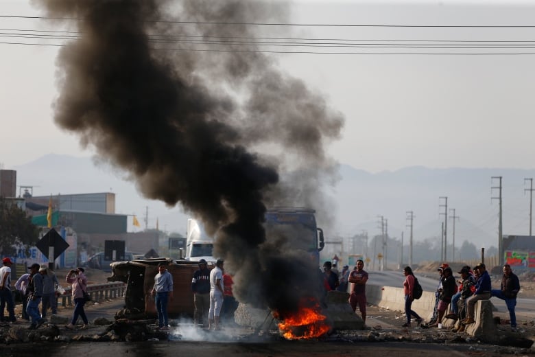 A column of black smoke rises from a fire, burning amid a gathering of protesters at a blockade of a highway.