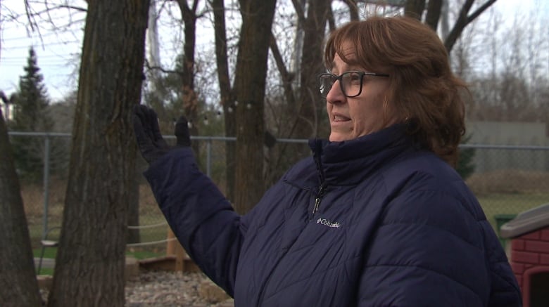 A woman with glasses and a dark blue winter jacket gestures at trees, standing in the yard of a daycare.