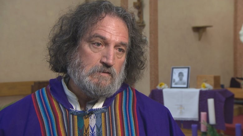 A priest wears a bright purple robe with multi-coloured Latin American stripes down the middle. Behind him, blurred, is a photo of Salomon Lázaro.