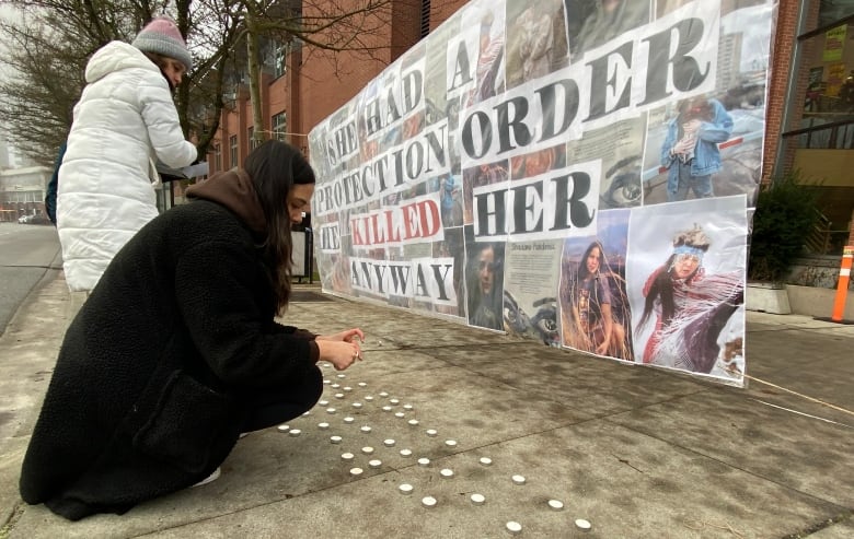 Two people lay votive candles in front of a banner reading 'She had a protection order; he killed her anyway.'