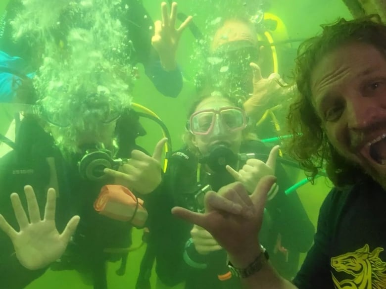 A man flashes a 'hang loose' sign with his hand in front of a round window peering into the ocean, where four scuba divers have gathered for a selfie. 