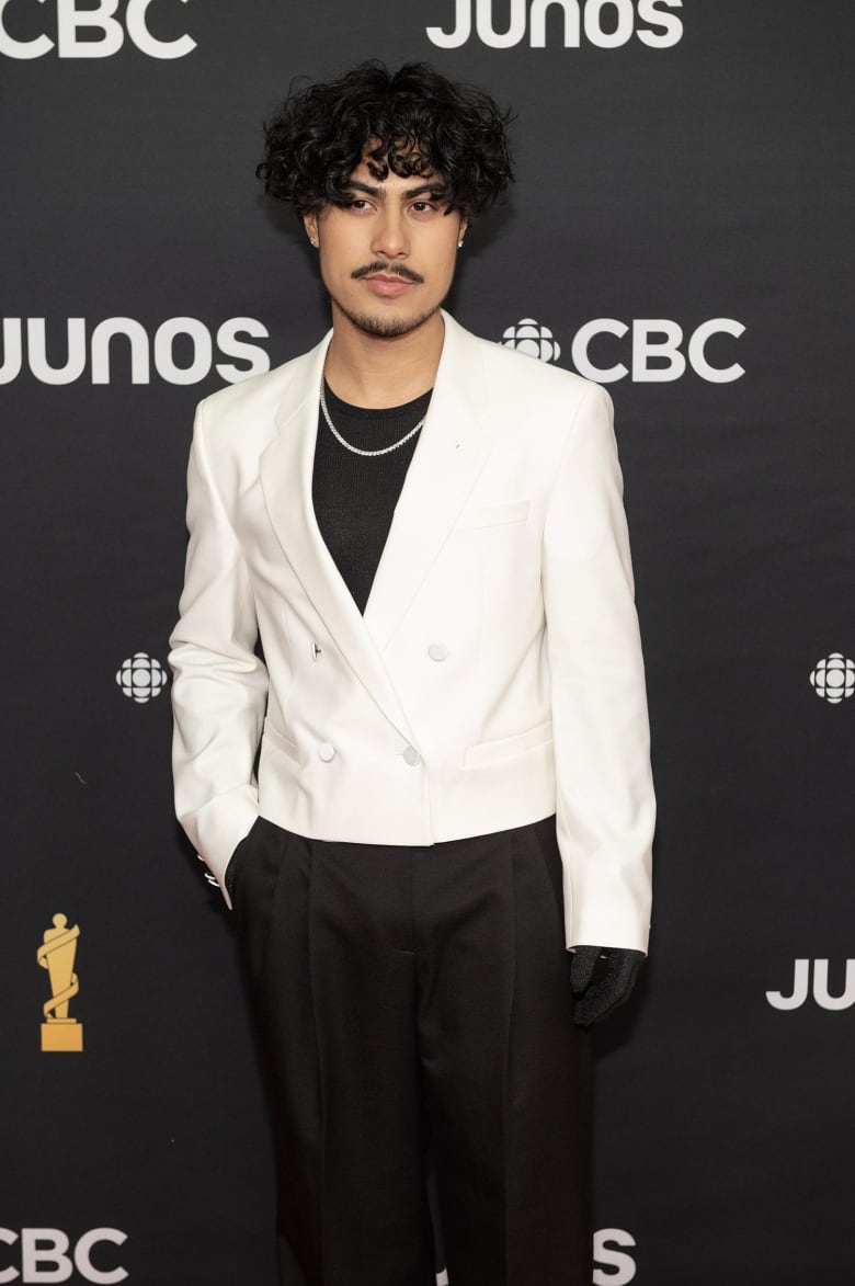 A man posing on the Junos red carpet wearing a cropped, double breasted white blazer, black pleated pants, a silver necklace and black gloves. 