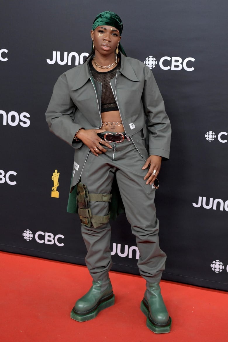 A woman posing on the Junos red carpet wearing a sheer cropped top, khaki-green jacket and pants, platform green boots, gold chains and mismatched earrings.  