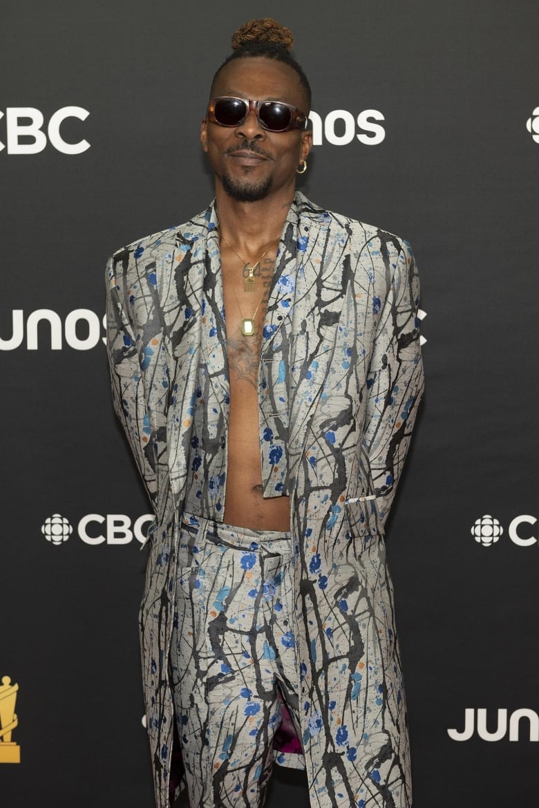 A man posing on the Junos red carpet wearing a three-piece suit without a shirt underneath, sunglasses and two layered gold necklaces. 