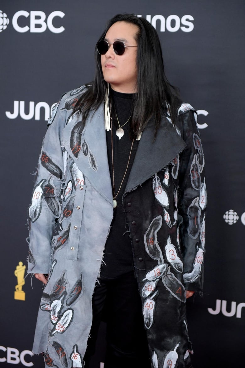 A man posing on the Junos red carpet in a grey, raw-edged coat featuring hand-stitched numbers on feathers. 