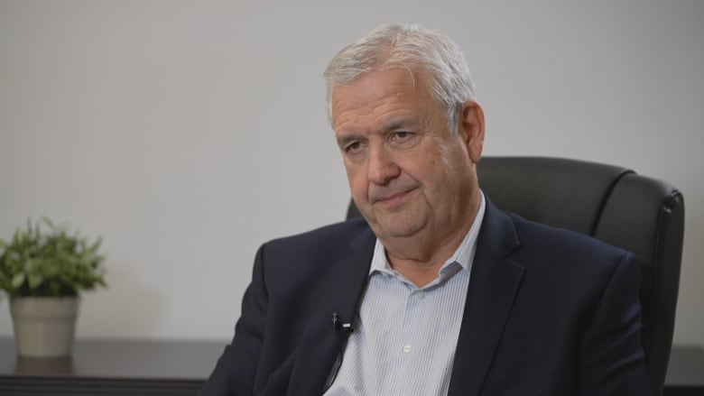 A man with white hair, wearing a blue and white striped shirt and a suit jacket, sits in an office chair.