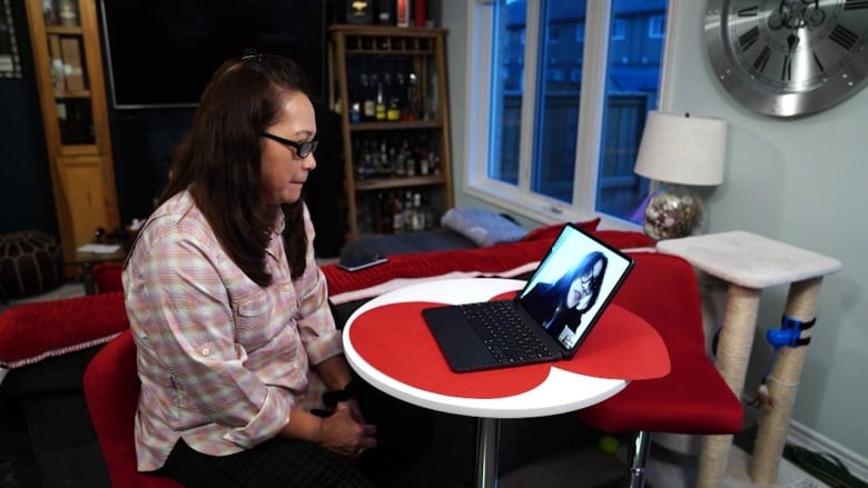 A woman with glasses, wearing a pink plaid shirt, sits at a small white table, with an open laptop on it. On the screen is a video call featuring another woman.