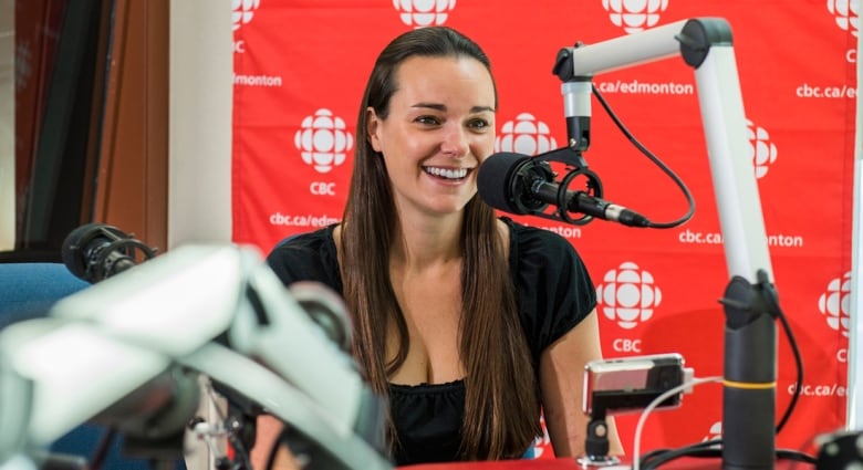 A woman smiles in front of a radio studio boom microphone in front of a red banner emblazoned with the logo of CBC Edmonton.