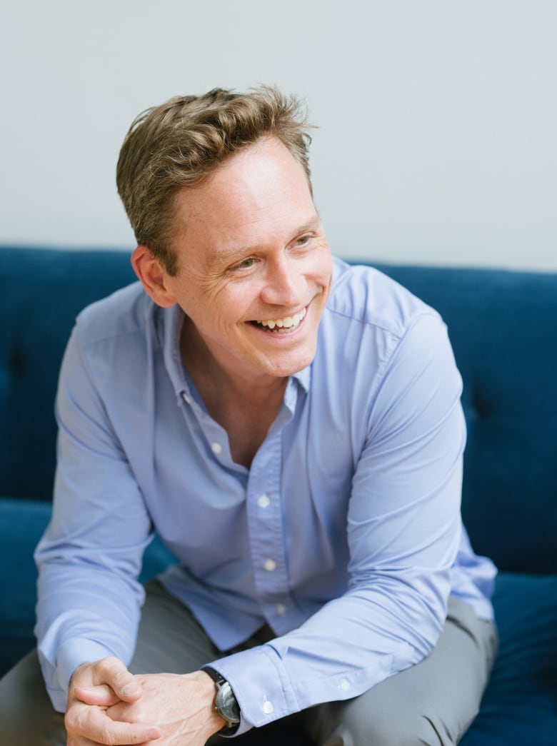 A man in a blue button-up shirt laughs while sitting on a sofa.