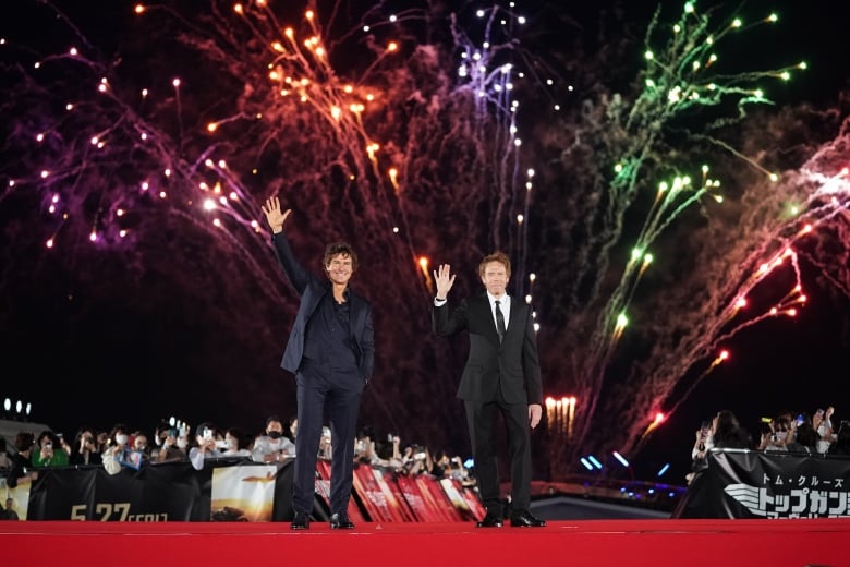 Two men stand waving on a red carpet. Behind them in the night sky are multicoloured fireworks. 