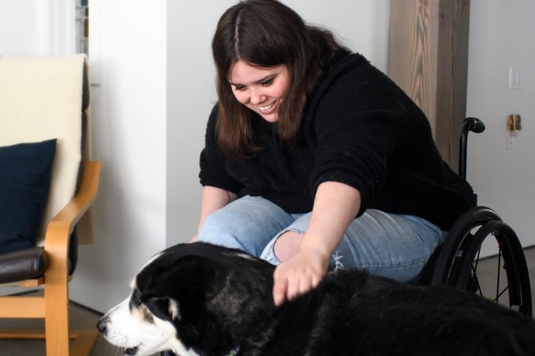 A woman in a wheelchair leans forward to pet her dog while smiling