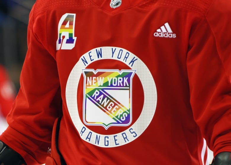 A red hockey jersey with a white circle surrounding a shield-shaped logo coloured with a rainbow. 