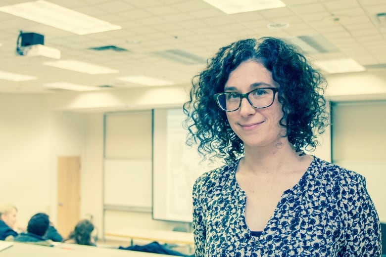 A woman with dark, curly hair and glasses stands in a classroom. 