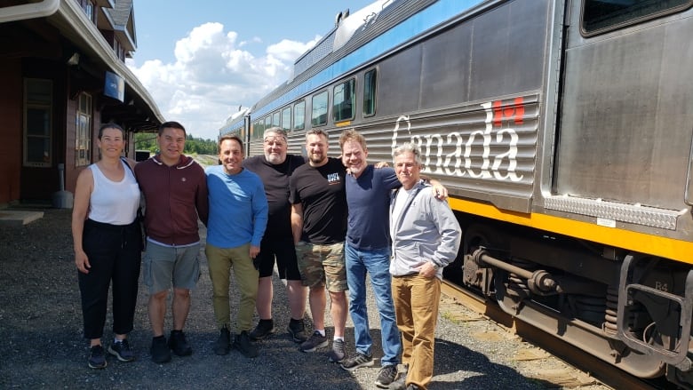 Seven people stand on the platform next to a stainless steel passenger car.