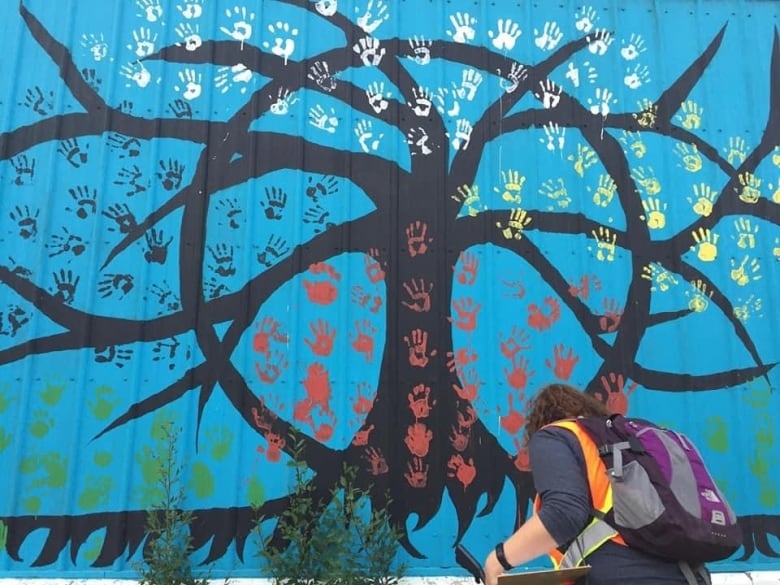 A woman reaches for something in the soil. She stands next to a mural of a tree covered in handprints.