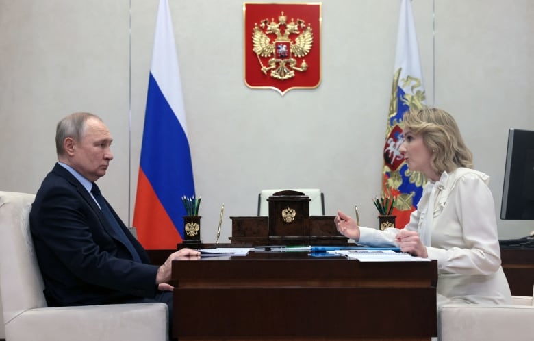 Woman in white sits across a brown desk from a man in a suit in front of flags