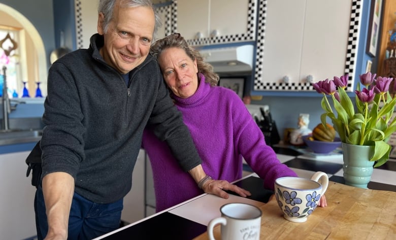 A man and woman stand in a kitchen. There are flowers and cups on the counter and she is leaning on him. 