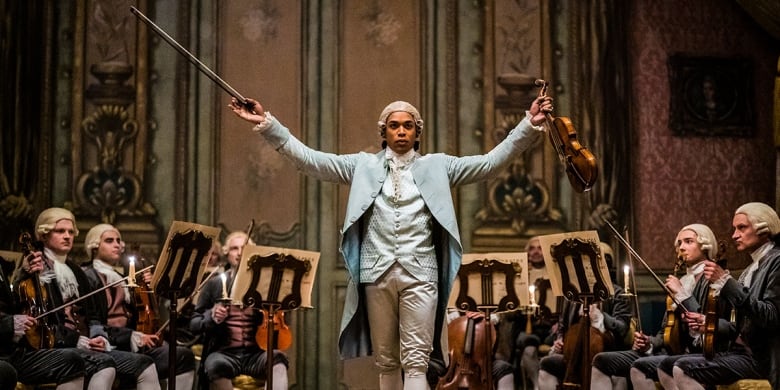 A Black man wearing an 18th century, French aristocratic outfit stands between a number of seated violinists, all of whom are white. He is holding a violin and bow in outstretched hands. 