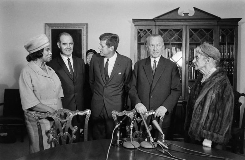 Five people pose in a black and white photograph. On the left is a woman in an old fashioned dress and hat, in the centre are three men, and on the right is an elderly woman in fur and a hat. They are standing in front of a table with three microphones pointed at them. 