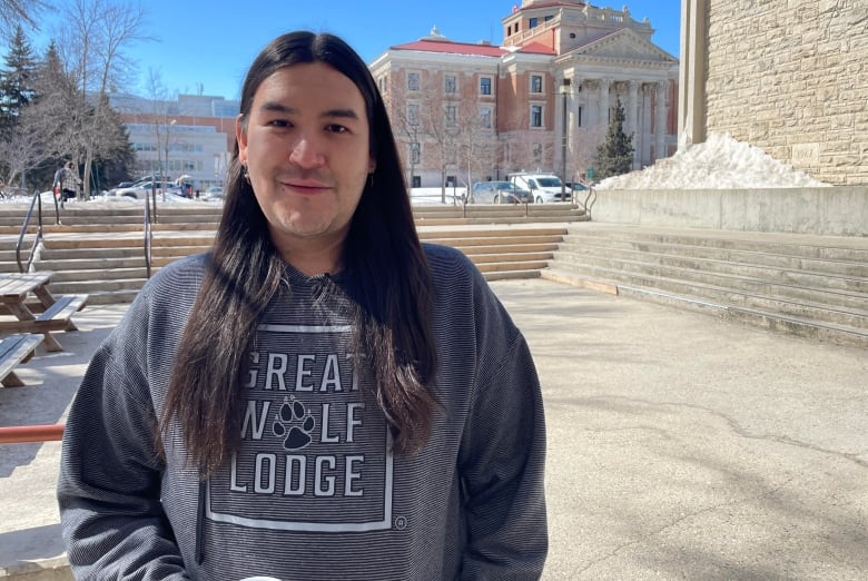 A young man stands outside of his university center and smiles at the camera