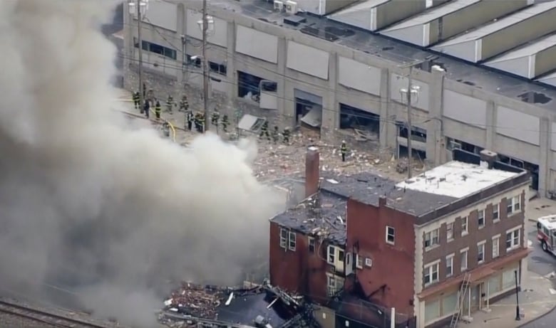 In an image shot from a height,  a collapsed building, a large plume of smoke, and about a dozen firefighters can be seen. 