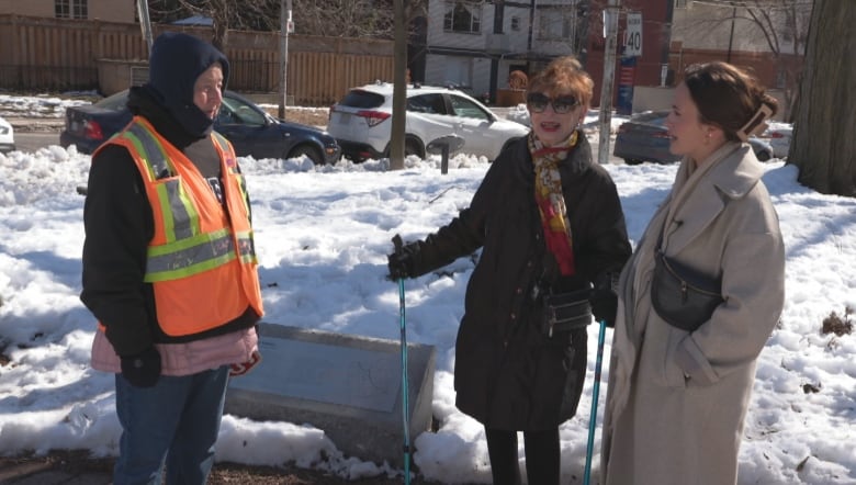 Three women are talking on a path at a park. One is wearing a crossing guard vest. Another is holding walking sticks.