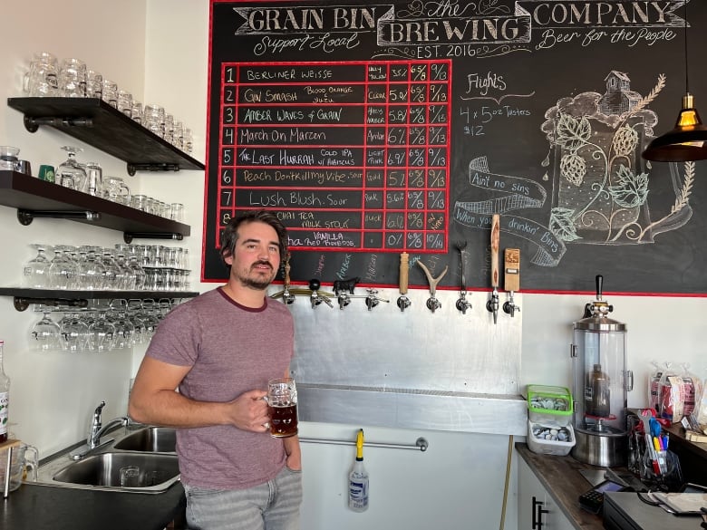 a man holds an amber beer behind a bar with tap[s and clean glasses.