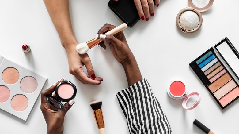 A woman's arm and hand on a cosmetics table surrounded by makeup, while another woman's hand brushes it with a cosmetic brush.