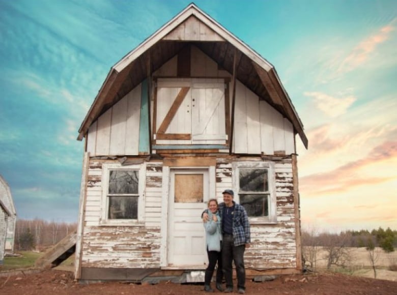 A man and woman stand outside a dilapidated farm building. 