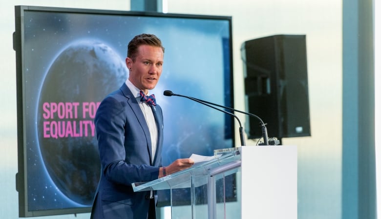 A man wearing a suit speaks while standing at a glass podium in front of a banner that reads sport for equality.