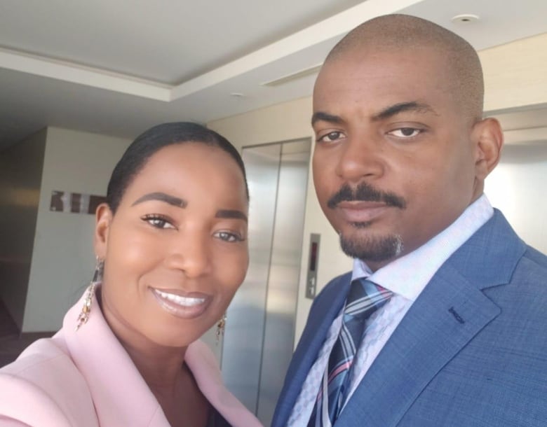 A man and a woman pose for a selfie in front of an elevator. The woman is wearing a light pink blazer, while the man is wearing a blue suit jacket, light blue checked shirt and blue plaid tie.