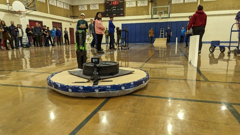 A circular unit with plastic film taped to the bottom and a leaf blower strapped to it sits at one edge of a gymnasium with a crowd looking onward at it