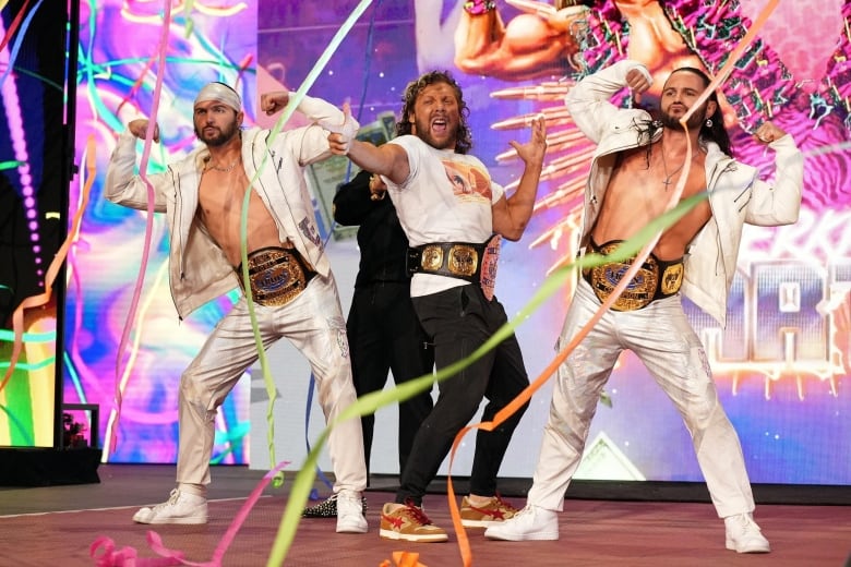 Three flamboyantly dressed pro wrestlers pose in front of the crowd.