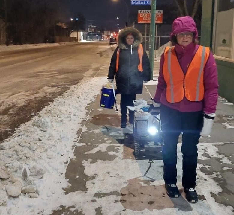 A woman in an orange safety vest pulls a cart loaded with first aid supplies and food on a snowy pavement.   