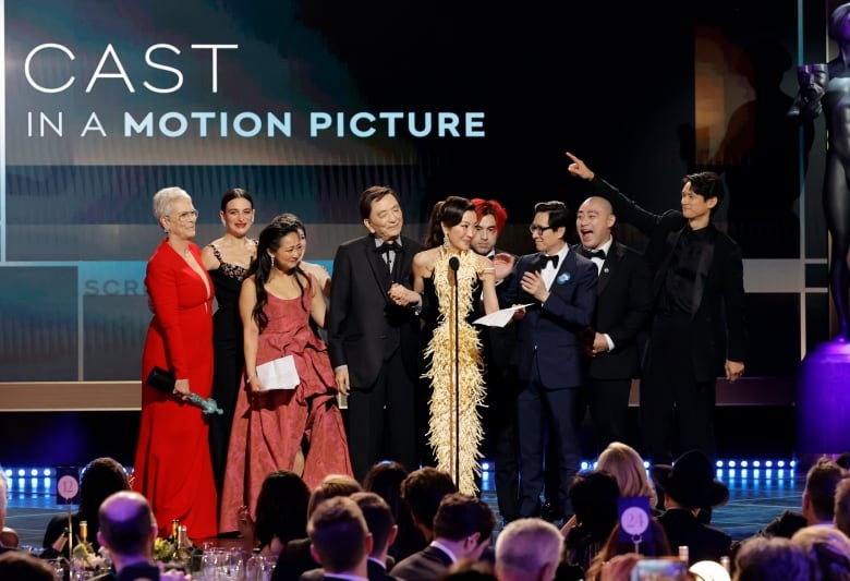 A group of ten stand onstage celebrating. Behind them the words "Cast in a motion picture" are visible. 