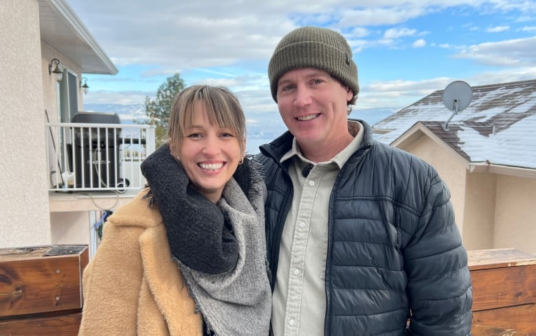 A couple wearing winter coats and scarves smile as they pose for a photo together on a balcony off their home.