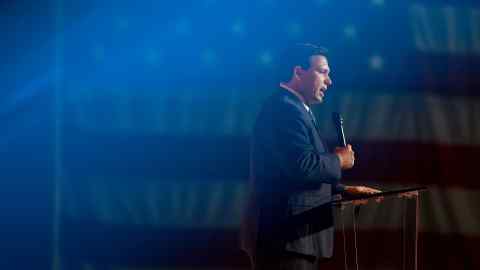 Ron DeSantis, the governor of Florida, standing at a podium with a microphone in his hand. A giant US flag hangs in the background